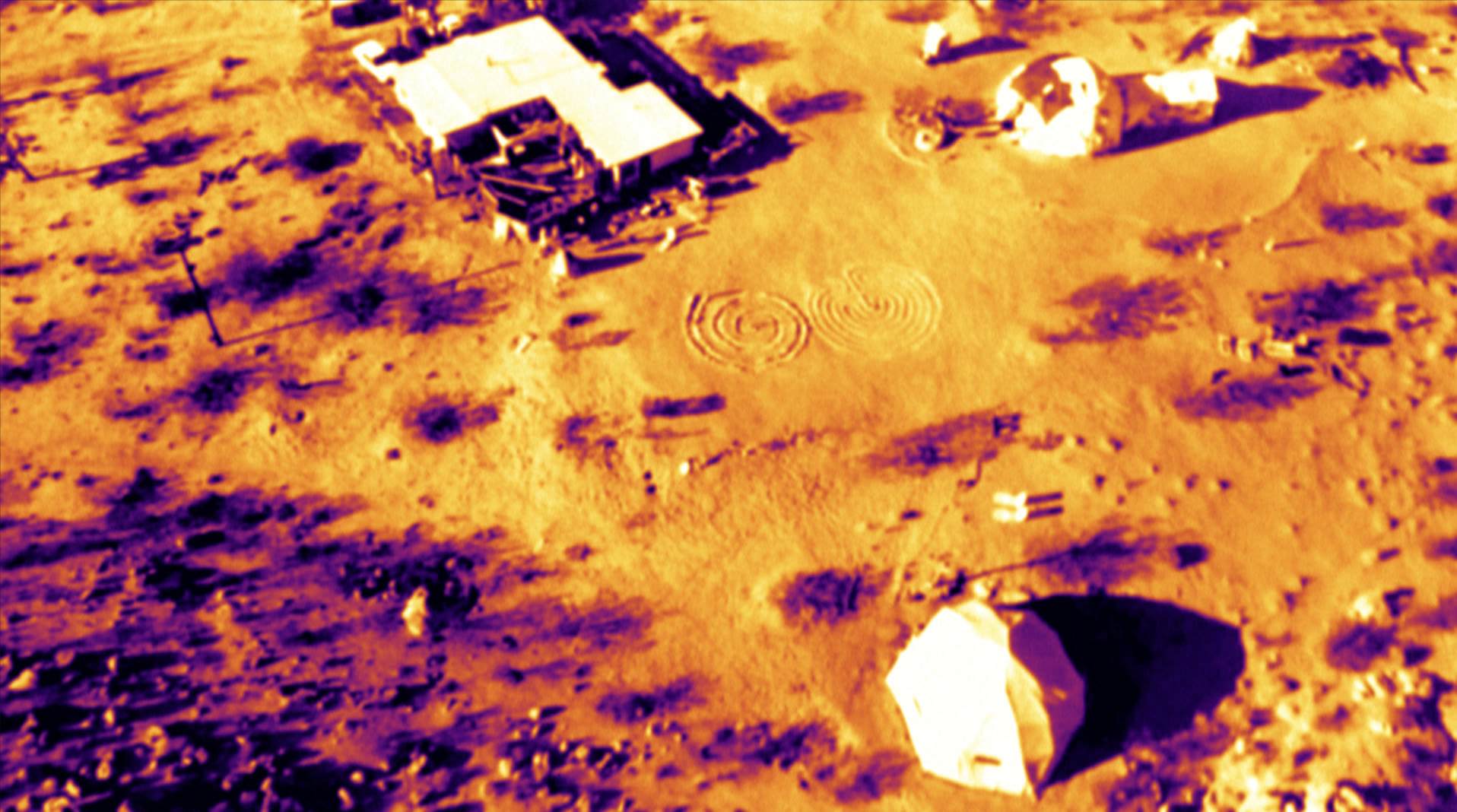 Thermal aerial imagery highlighting structures, vehicles, and terrain features.
